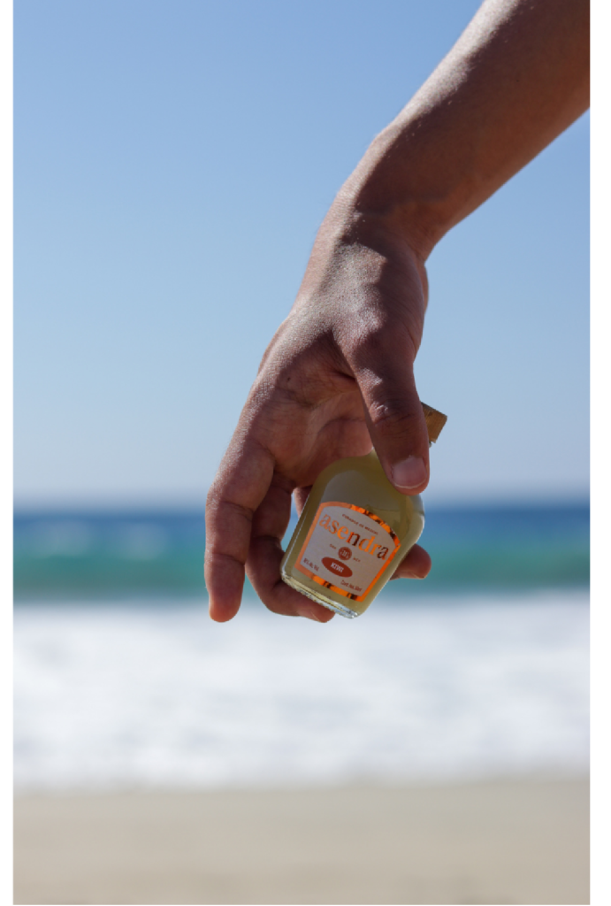 mano sosteniendo una botella pequeña del mezcal asentar con la playa de fondo