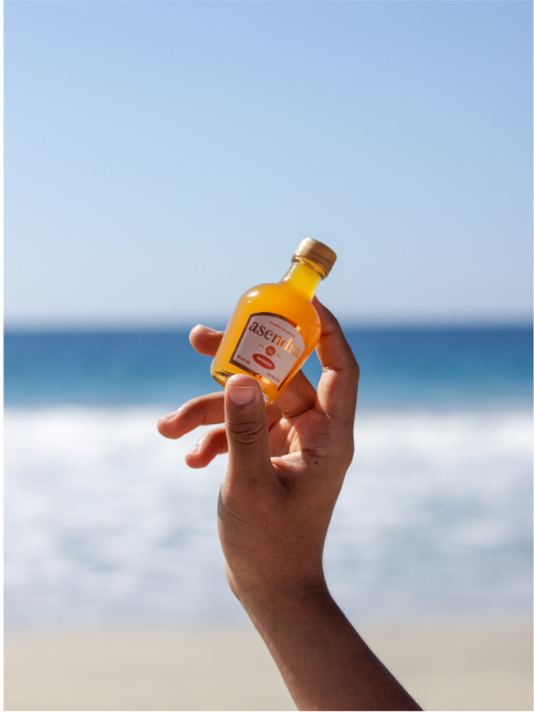 Mano sosteniendo una botella pequeña de Mezcal Asendra con la playa de fondo